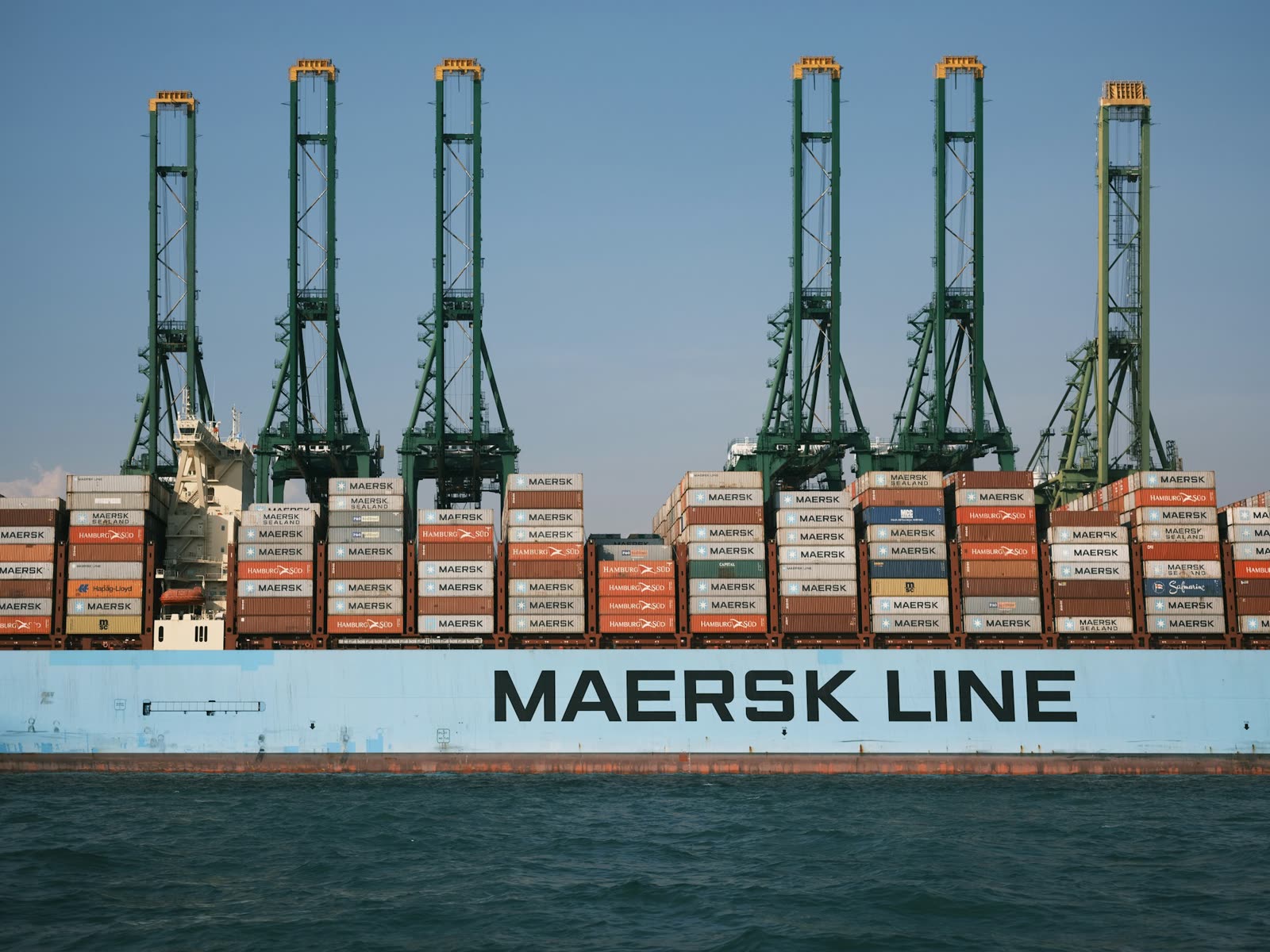 The hull of a container vessel marked Maersk Line, with stacked containers and green cranes behind it.