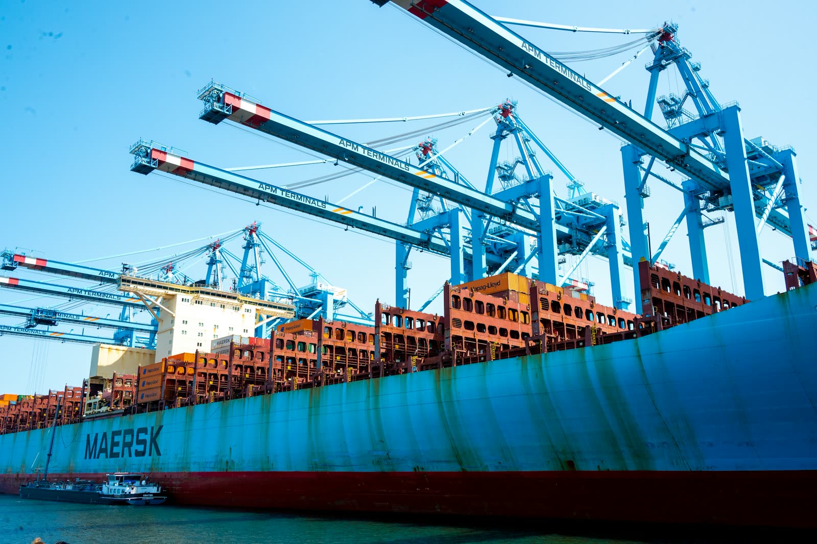 The light-blue hull of a container vessel with the Maersk name, alongside under blue quay cranes.