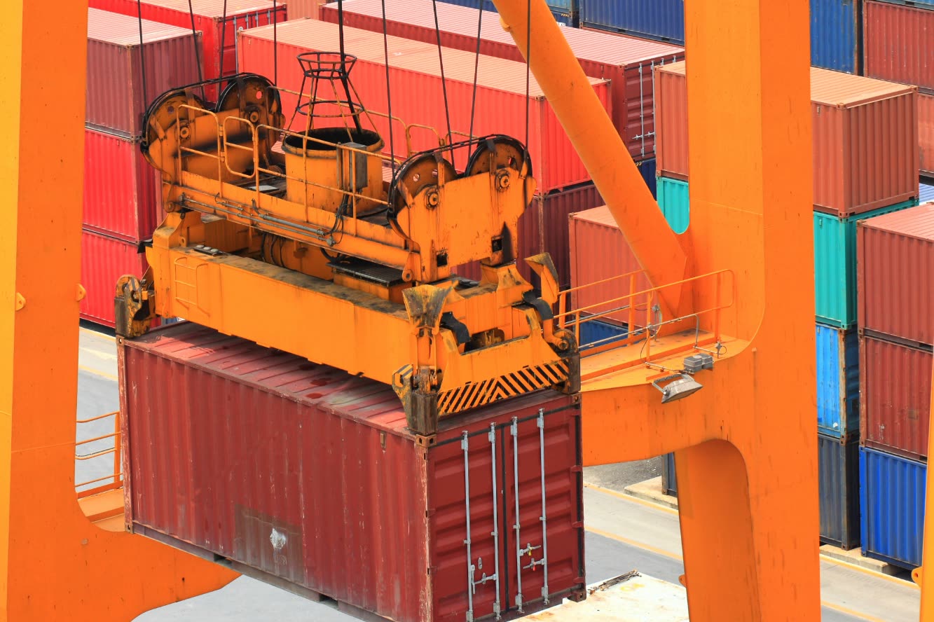 A crane spreader lifting a container, with stacked containers behind it.